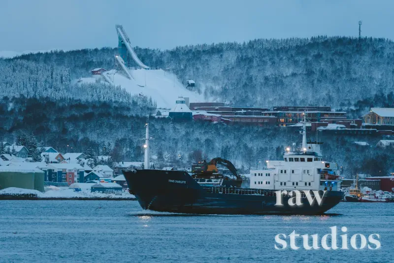 Et stort, mørkt skip seiler gjennom snødekt vann i et maritimt landskap med fjell og en hoppbakke. Omgivelsene består av en bebygd kystlinje under en blålig vinterhimmel med en rolig stemning.
