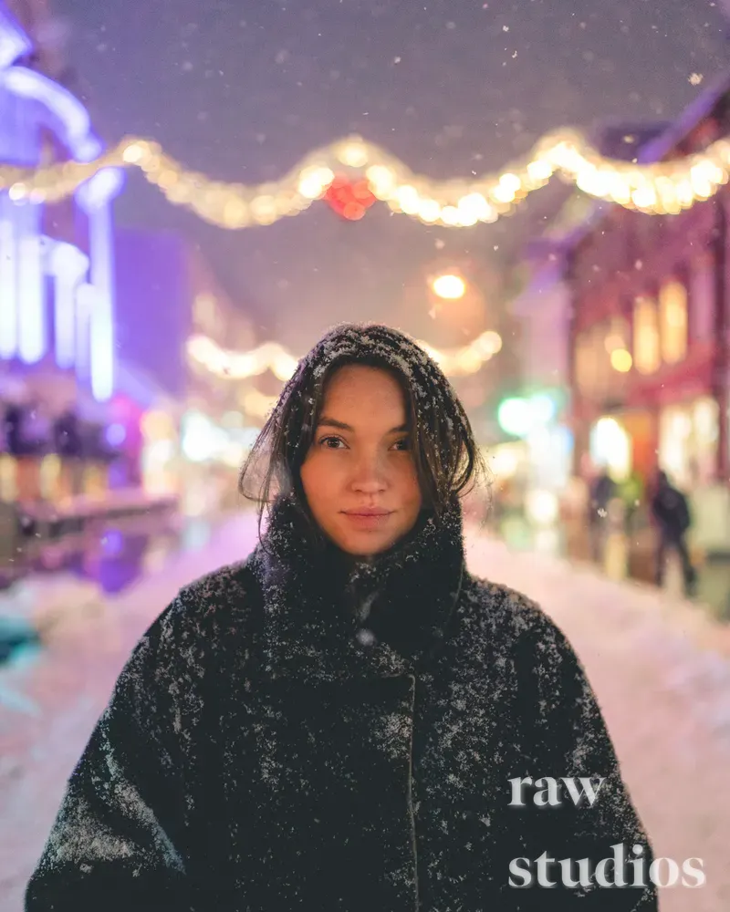 Portrett av en kvinne i mørke vinterklær på en snødekt gate under et lett snøfall. Bakgrunnen har varme lyslenker og glødende dekorasjoner som skaper en stemningsfull atmosfære med innslag av blått og lilla lys.