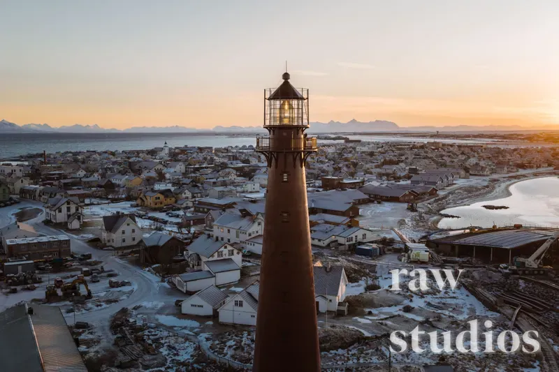 Et mørkt fyrtårn rager over en snødekt kystlandsby ved havet under en varm solnedgang. Landskapet preges av små hus, fjell og vann i et rolig vinterlandskap med gyllent lys.