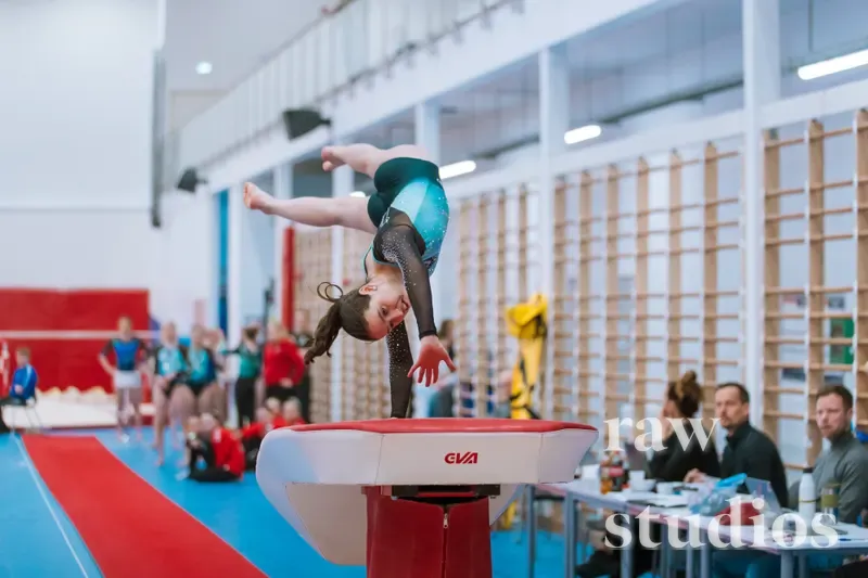 En kvinnelig gymnast utfører et hopp over en hopphest i en lys turnhall eller idrettshall. Bakgrunnen viser klatrevegger av tre og andre utøvere under kunstig belysning, med farger som rødt, blått og grønt på treningsmattene.
