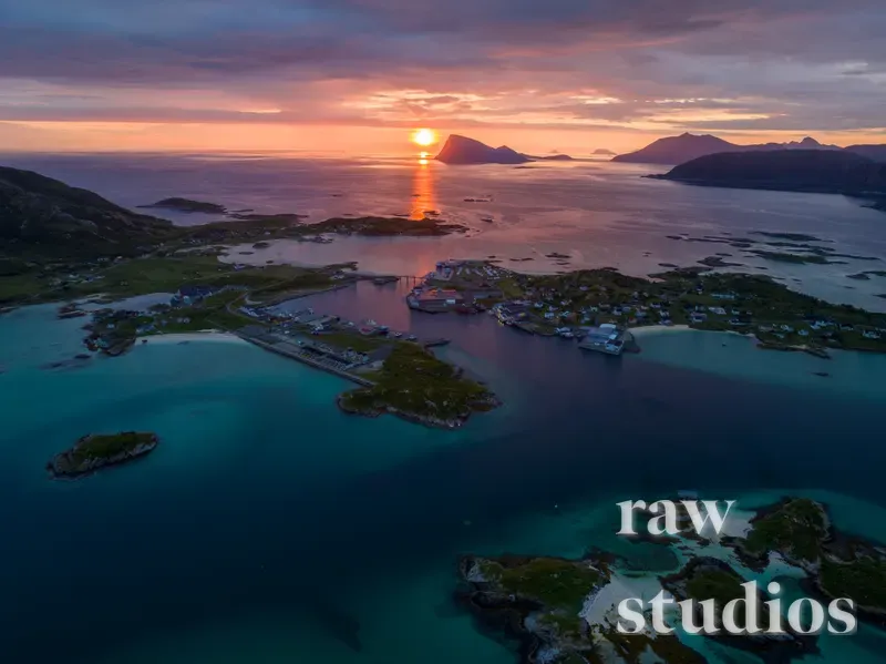 Droneperspektiv av et kystlandskap med øyer, fjell og små bosetninger ved havet under en varm solnedgang. Vannet og sjøen viser nyanser av blått og turkis mot en himmel i oransje og rosa toner.