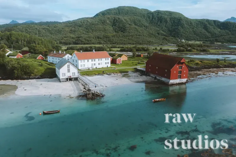 Luftfoto av et maritimt kystlandskap med hvite og røde trehus ved en strand under grønne fjell. Det klare, turkise havet omkranser bebyggelsen i sommerlig dagslys, med små båter som ligger i vannet.