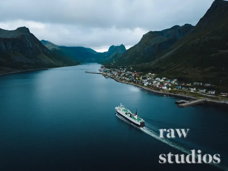 Et dronebilde viser et skip som seiler gjennom en fjord mellom bratte fjell og en liten kystlandsby. Det maritime landskapet preges av blått vann, mørke fjellsider og en overskyet himmel som skaper en rolig stemning.