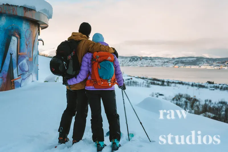 To personer med ryggsekker står i et snødekt vinterlandskap og ser utover en fjord eller kystlinje mellom fjellene. Bildet viser friluftsliv ved sjøen under en lys, overskyet himmel.
