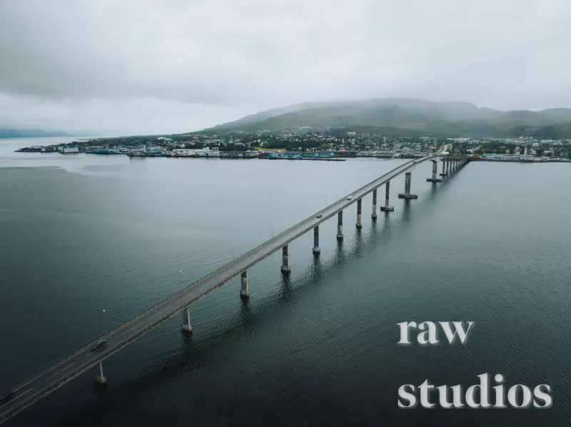 Et dronebilde av en lang bro som krysser sjøen mot en kystby ved foten av fjellene. Landskapet viser en havn og maritim infrastruktur under en overskyet himmel med dempede farger og en rolig stemning.