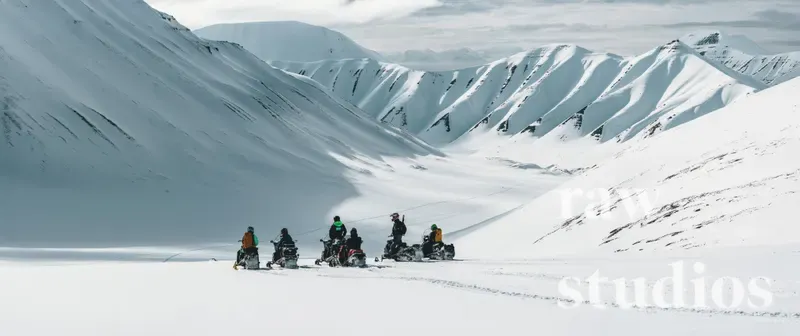 En gruppe mennesker på snøskutere kjører gjennom et vidstrakt og snødekt fjellandskap under en overskyet himmel. Bildet viser vinterlig friluftsliv i alpint terreng med hvite fjellsider og dempet lys.