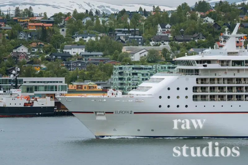 Et stort hvitt cruiseskip seiler gjennom en rolig fjord forbi en kystby med fargerike hus og snødekte fjell i bakgrunnen. Landskapet preges av grønne trær, blått vann og sommerlig dagslys.