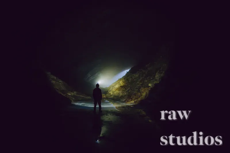 En person står som en silhuett i en mørk grotte eller tunnel ved et stille vann. Dramatisk lys fra en åpning skaper en mystisk stemning med sterke kontraster og refleksjoner i vannflaten.