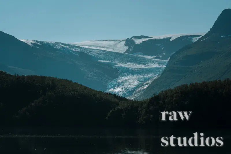 Et majestetisk fjellandskap med en lys blå isbre som renner mellom mørke fjellsider mot en skogkledd dal. Forgrunnen viser tett skog og stille vann under en klar, blå himmel i kaldt dagslys.