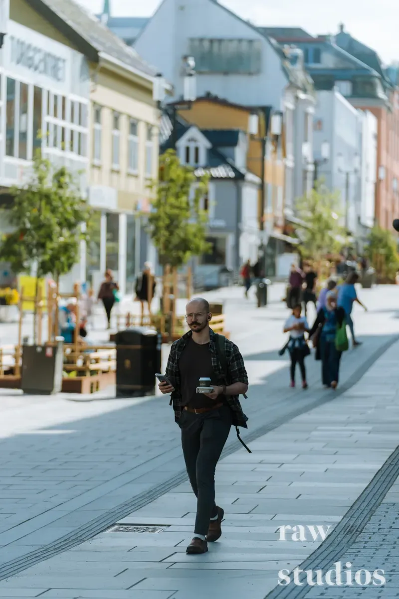 En skjeggete mann går gjennom en travel gågate i et bysentrum med kaffekopp og mobiltelefon i hendene. Omgivelsene består av bygninger, trær og andre fotgjengere under dagslys i sommerhalvåret.