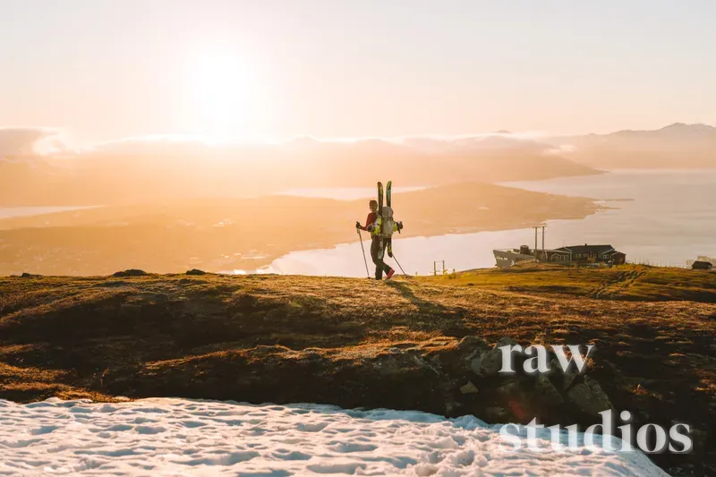 En person med ski på ryggen vandrer gjennom et fjellandskap ved kysten under en varm solnedgang. Landskapet består av fjorder, fjell og rester av snø i et gyllent lys.