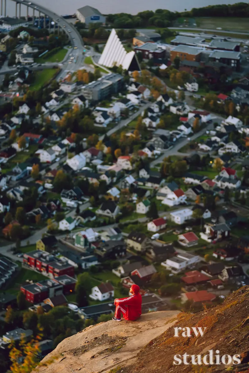 En person i rød jakke sitter på en klippe og skuer ut over et boligområde med hus, veier og grøntområder. Dette panoramaet viser bebyggelse og infrastruktur sett fra høyden i et rolig høstlandskap.